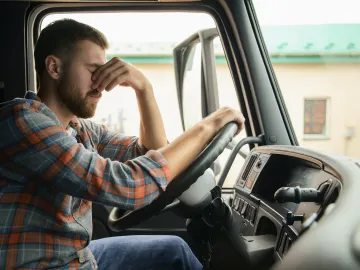 Tired truck driver sitting in the cabin with eyes closed and hand on his forehead, showing exhaustion while holding the steering wheel logo